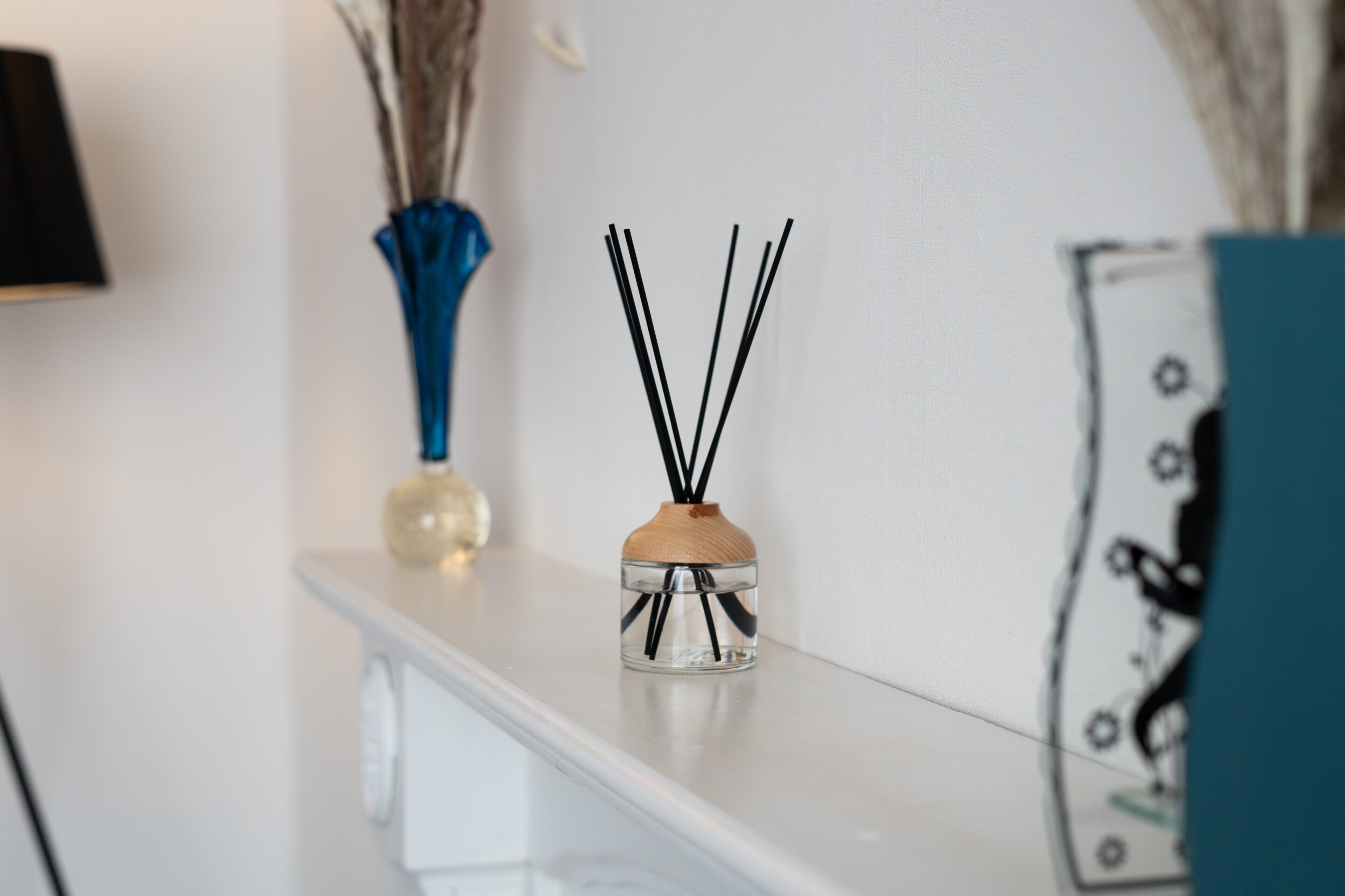 A clear glass diffuser with black reeds on a white shelf, a blue vase with dried plants, and a decorative item with a blue and white pattern.