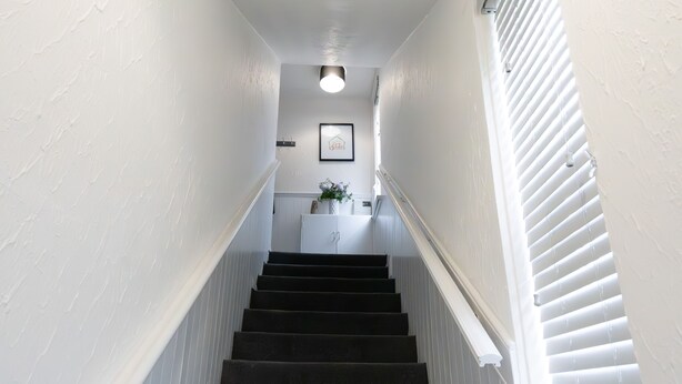 A staircase with white walls and a dark carpeted floor, leading to a landing with a white cabinet and a framed picture. - 2-bed Flat, Wishaw, Free Parking, Family-friendly (Wishaw)