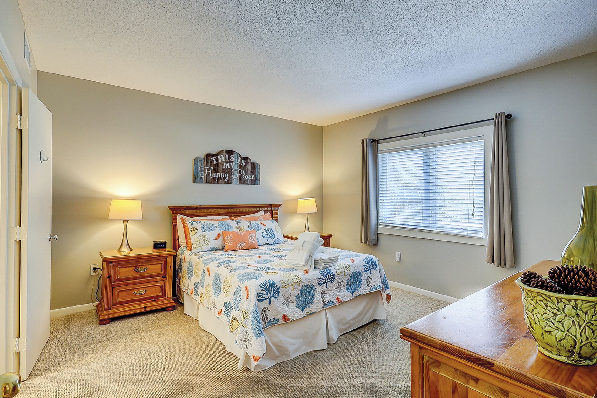 A bedroom with a bed, two bedside tables, a nightstand, a window with blinds, a decorative sign, and a vase with pinecones.