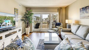 A living room with a sofa, a coffee table, a TV, a plant, and a balcony with a view of palm trees and a pool. - Sunset Rentals | Shorewood 134 (Hilton Head Island)