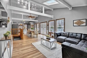 Un salón espacioso con techo alto, pisos de madera, un sofá de cuero negro y un comedor con mesa y sillas de madera.