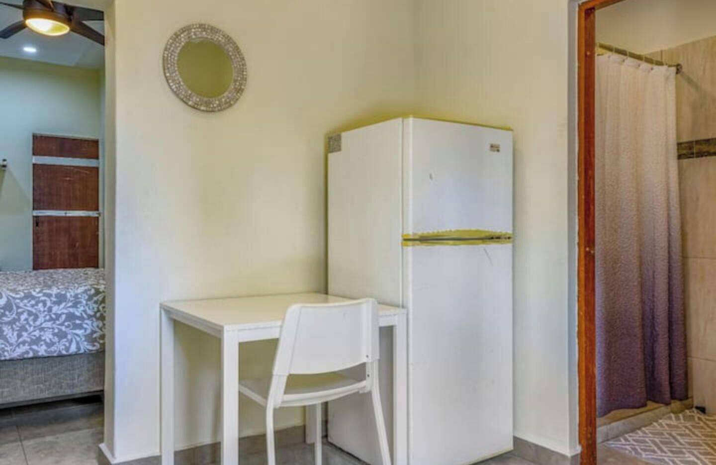 A small room with a white desk and chair, a white refrigerator, a mirror on the wall, and a bed with a patterned blanket.