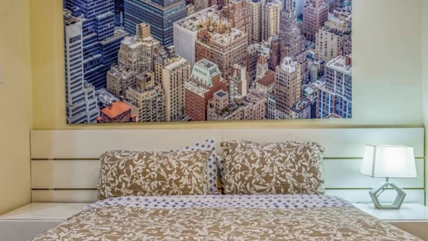 A hotel room with a large cityscape painting on the wall, a bed with patterned bedding, and a nightstand with a lamp.