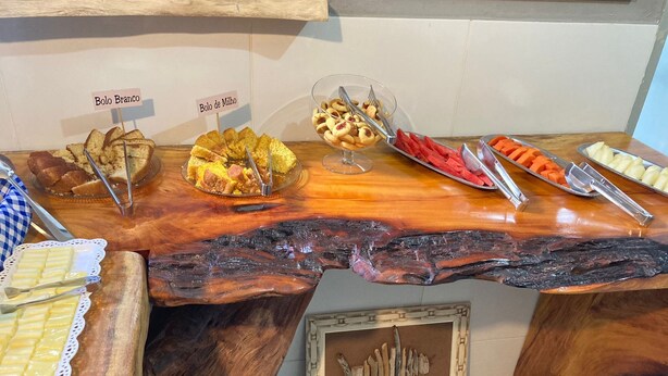 Een buffettafel met borden die gelabeld zijn, zoals Bolo Branco en Bolo de Milho, plus verschillende soorten fruit en een decoratief houten object.