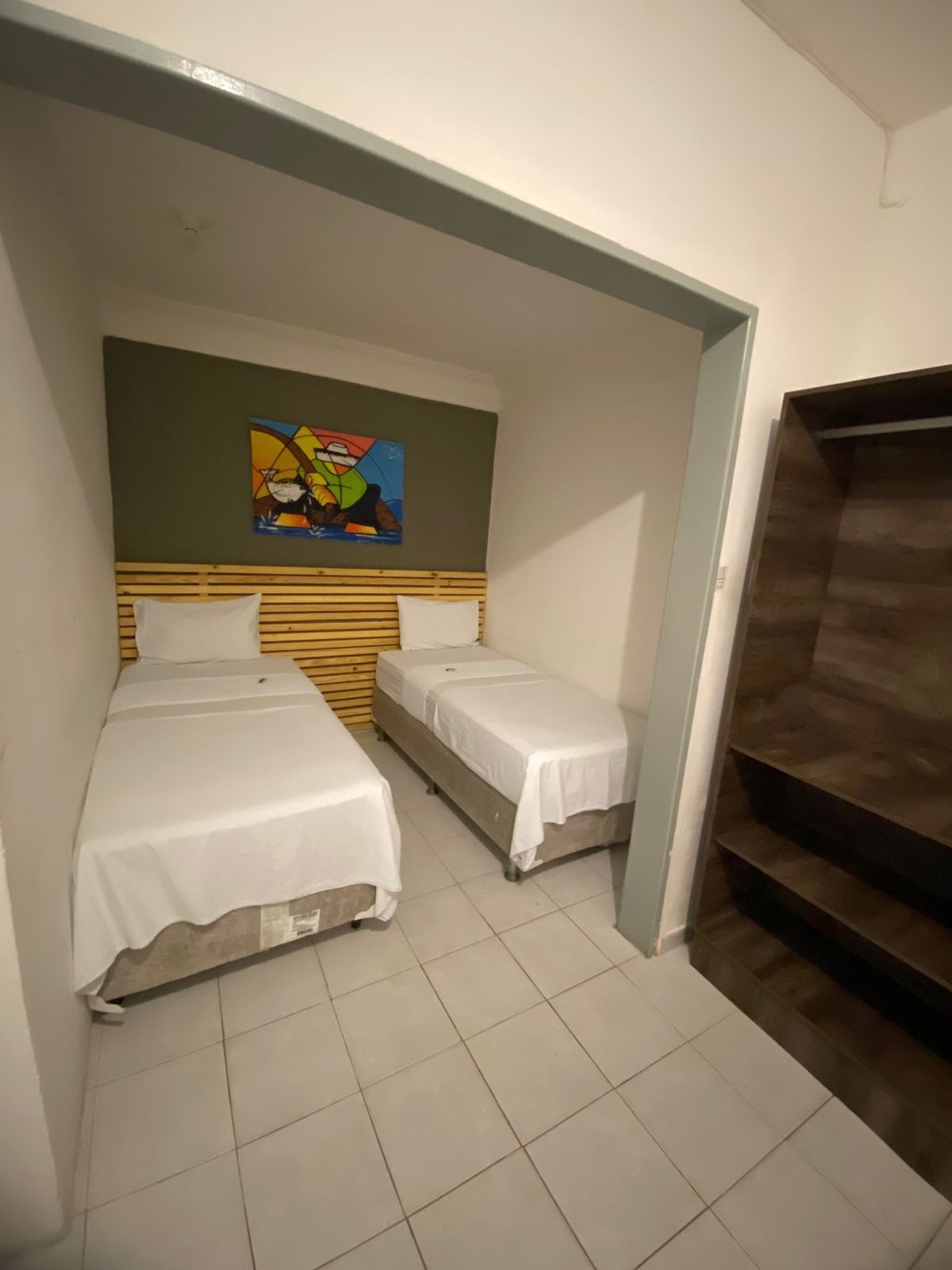 Una habitación pequeña y funcional con dos camas individuales, cabecero de madera, un cuadro abstracto de colores y un armario de madera.