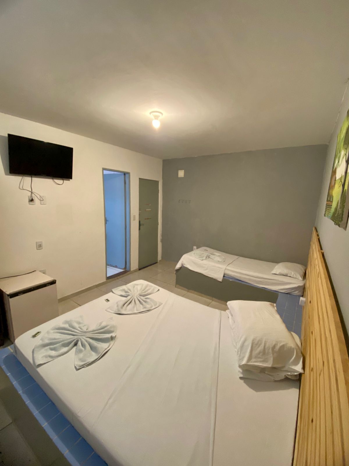 Habitación de hotel con una cama individual, un pequeño escritorio, un televisor montado en la pared y una puerta que da a otra habitación.