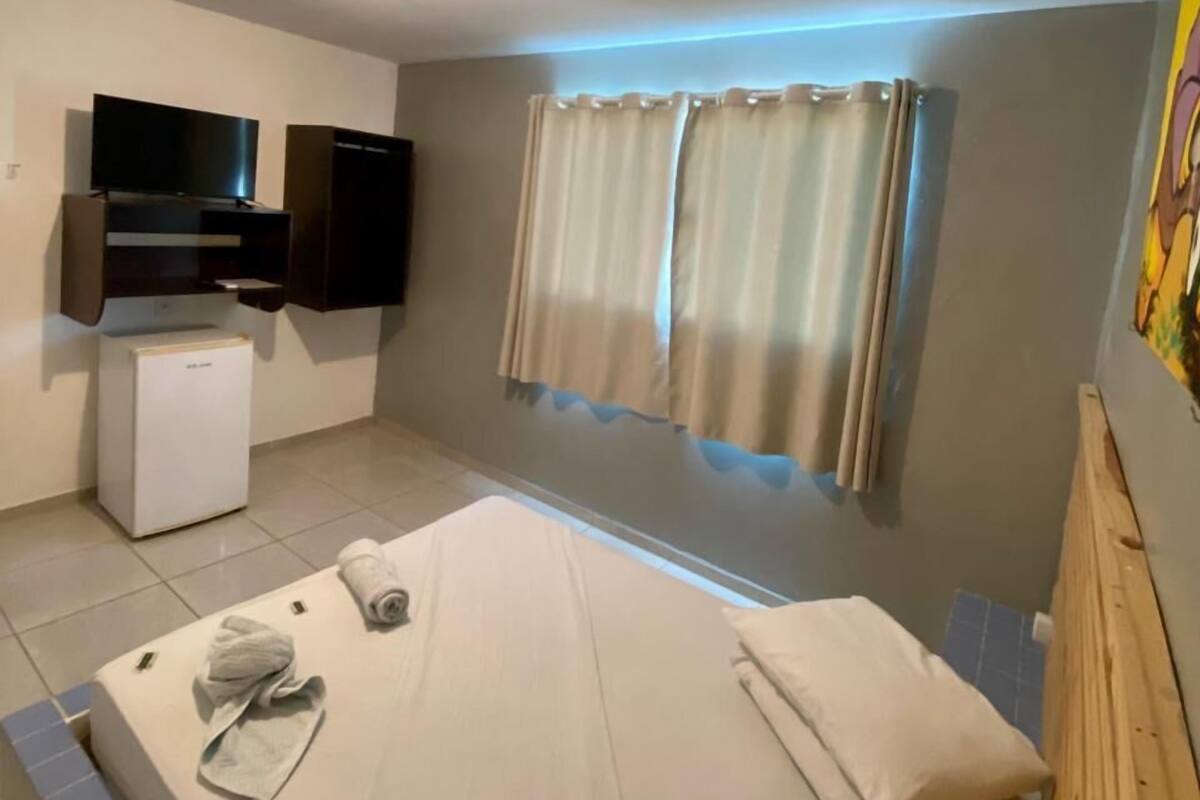 A bedroom with a bed, a TV, a small fridge, and a window with curtains.