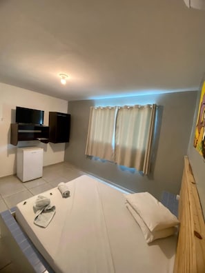 A bedroom with a bed, a TV, a small fridge, and a window with curtains. - Pousada Nascer do Sol  (João Pessoa)