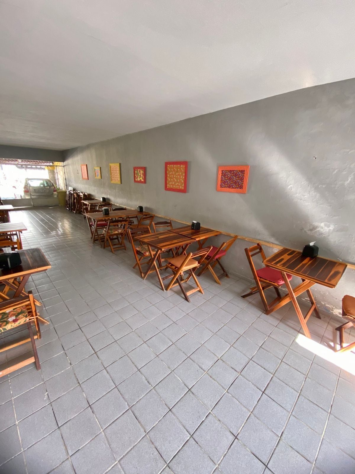 A spacious room with multiple wooden tables and chairs arranged in rows, a tiled floor, and a plain wall with framed artwork.