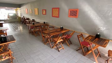 Un'ampia stanza con diverse tavoli e sedie in legno disposti in file, pavimento piastrellato e parete spoglia con opere d'arte incorniciate.