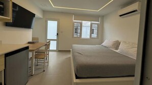 A compact bedroom with a bed, a desk, and a chair. There is a window with blinds, a door, and an air conditioning unit on the wall. - Beach House Walk to The Beach #8 (San Juan)
