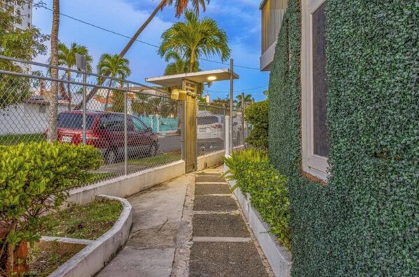 A pathway leading to a building with a green wall, a fenced parking area, and a palm tree in the background.