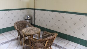 Un angolo con due poltrone in vimini, un tavolino, un cestino metallico e una parete piastrellata con bordo verde.