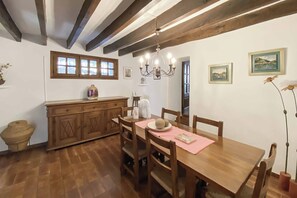 A dining area with a wooden table, chairs, and a chandelier. There is a window with multiple panes and a wooden cabinet with a vase on top.