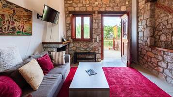 A cozy living room with a stone wall, a flat-screen TV mounted on the wall, a grey sofa with red and white cushions, a white coffee table, and a red carpet.