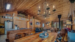 Interior rústico de cabaña con mesa de madera, bancos, una araña y una zona de cocina con refrigerador y armarios.