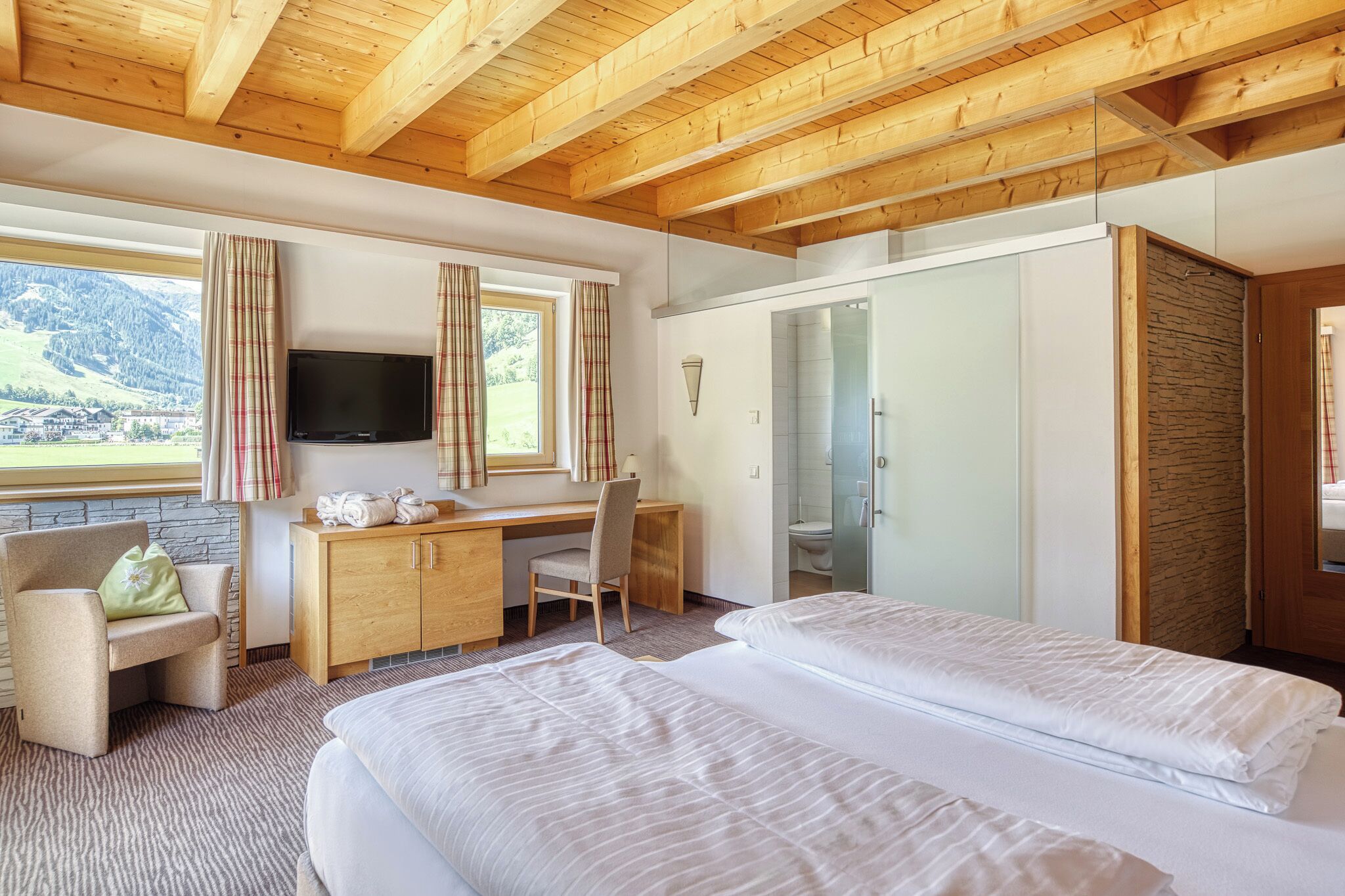 A hotel room with a wooden ceiling, a bed, a desk with a chair, a TV, and a view of mountains.