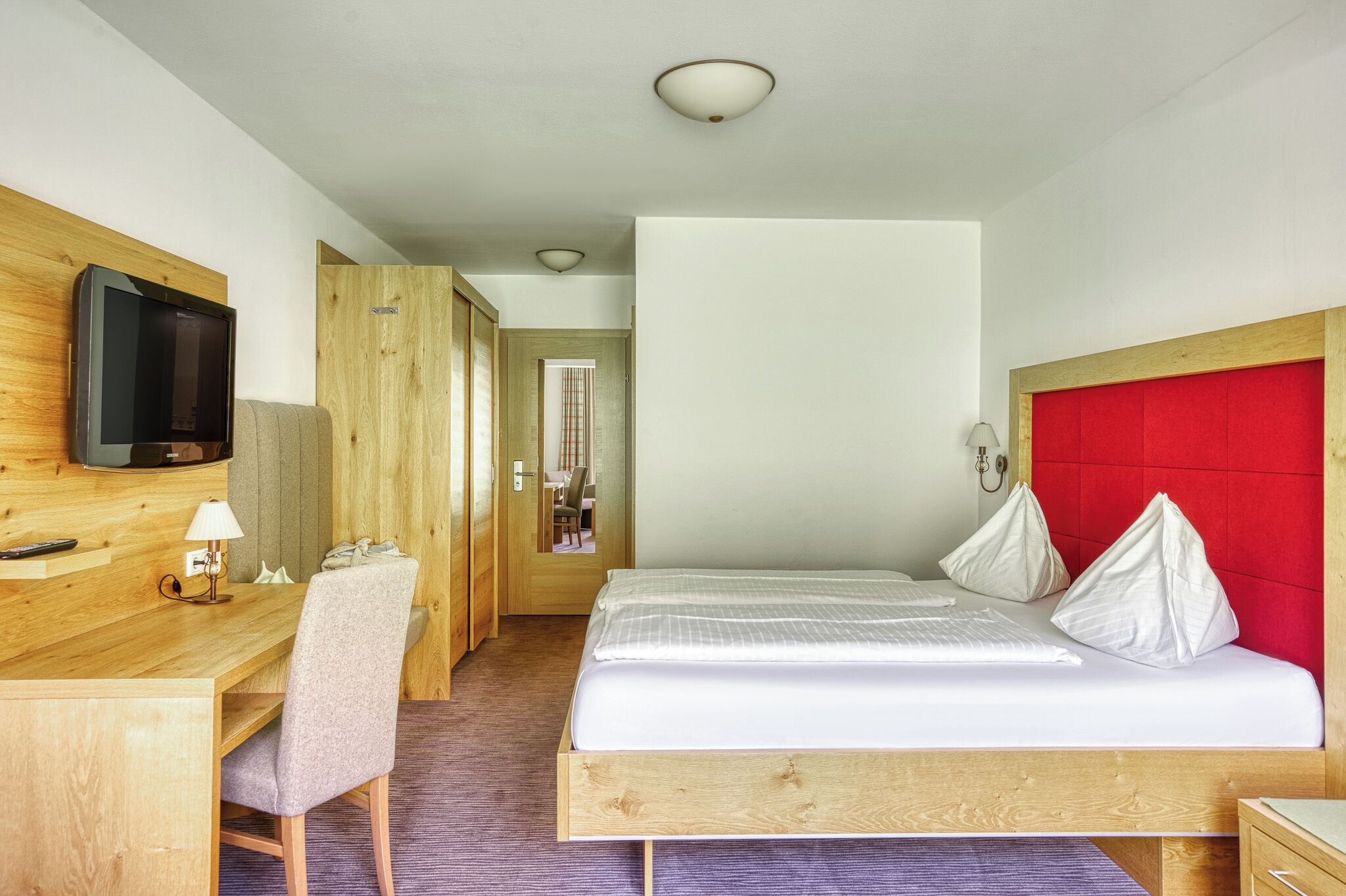 A hotel room with a bed, a desk with a chair, a television, and a red headboard.