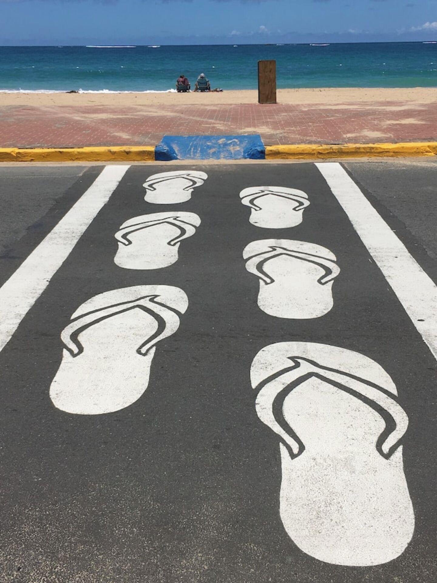 A parking area with footprints painted on the ground, leading to a beach with people sitting on the sand and boats in the water.