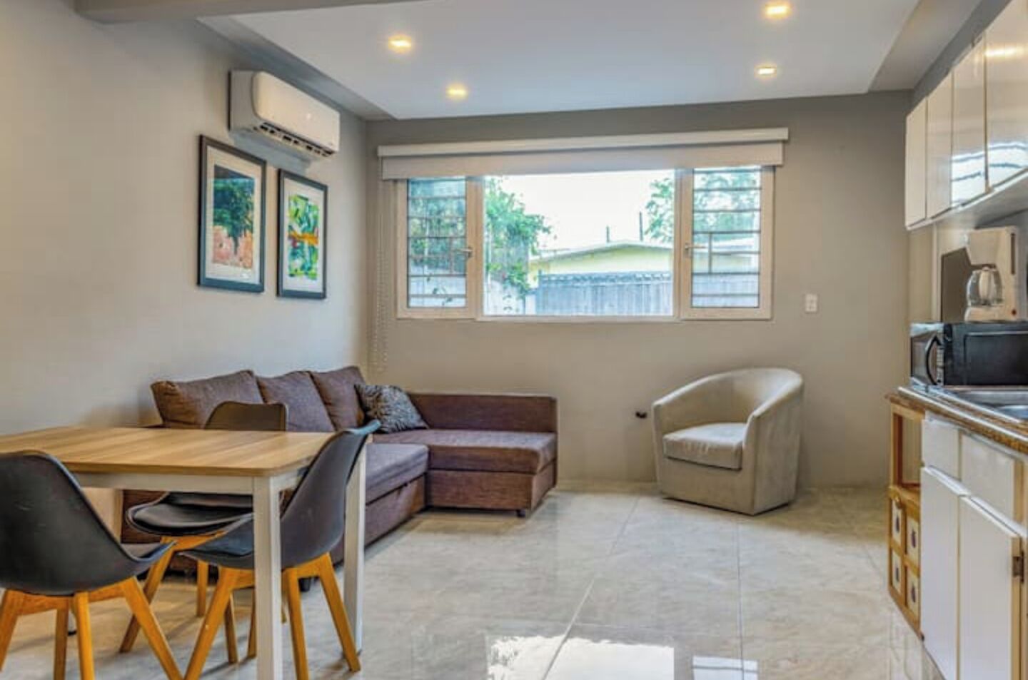 A modern living room with a sofa, a dining table with chairs, a kitchen area with cabinets, and large windows providing natural light.
