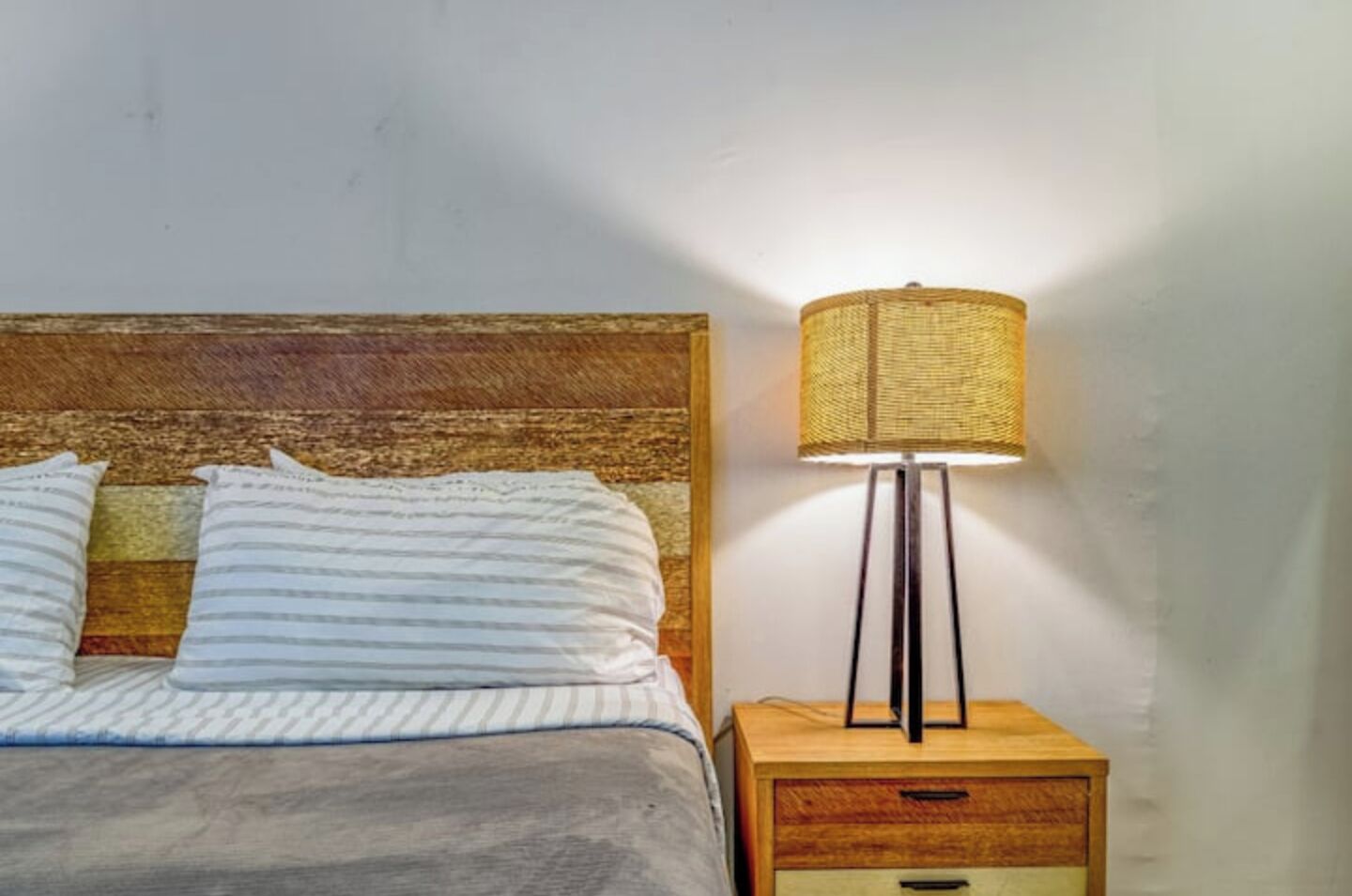 A bed with striped pillows and a wooden headboard, a bedside table with a lamp, and a small drawer.