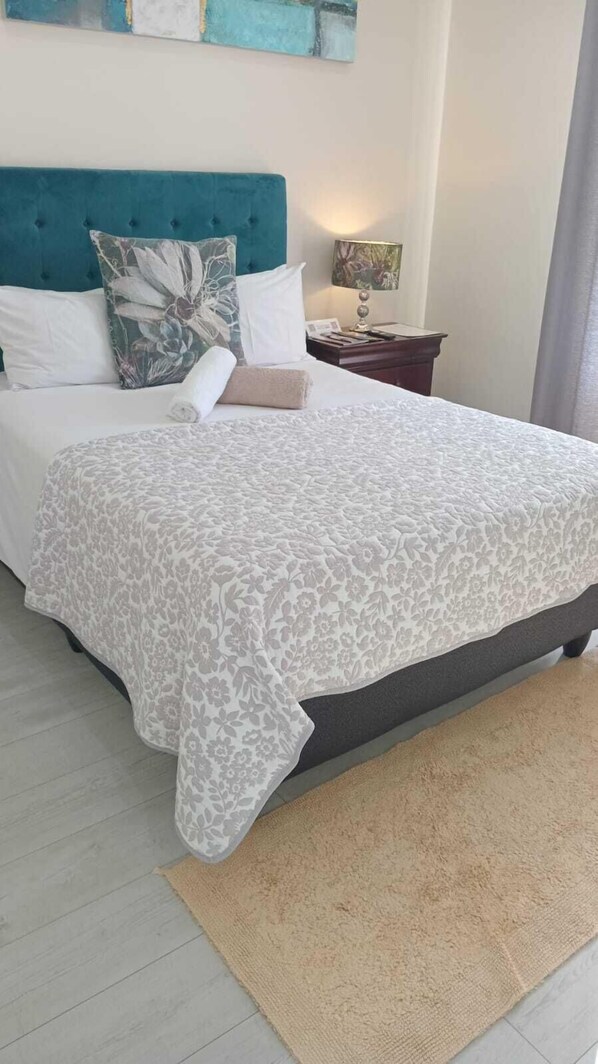 A neatly made bed with a floral-patterned bedspread, a teal headboard, and a bedside table with a lamp. - LND GUEST HOUSE (Cape Town)