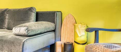 A room with a grey sofa, a wooden surfboard, a yellow trash bin, and a patterned chair.