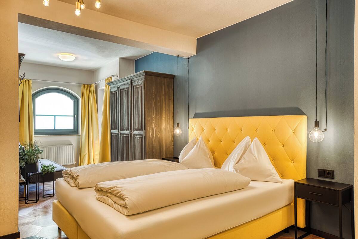 Une chambre d’hôtel avec une tête de lit jaune, un lit avec du linge de lit blanc, une table de chevet sombre et une fenêtre avec des rideaux jaunes.