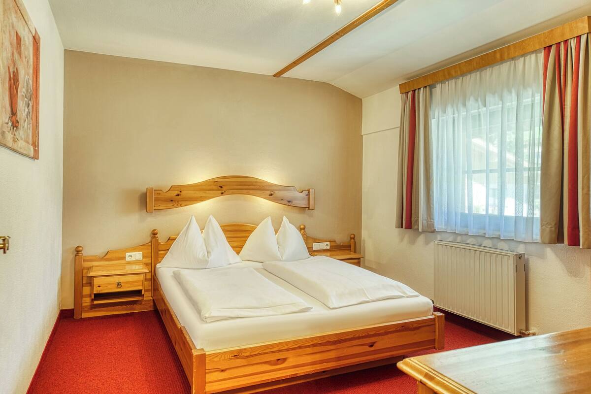 Une chambre d’hôtel avec un lit en bois, une table de chevet, une commode, une fenêtre avec des rideaux et un radiateur.