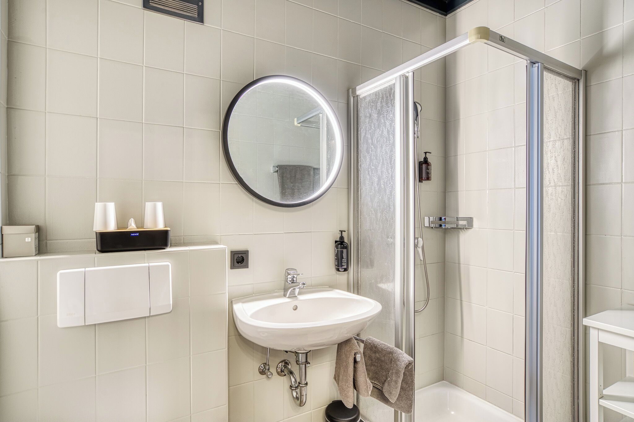 Un baño moderno con un espejo redondo, un lavamanos, una ducha y un perchero para toallas.
