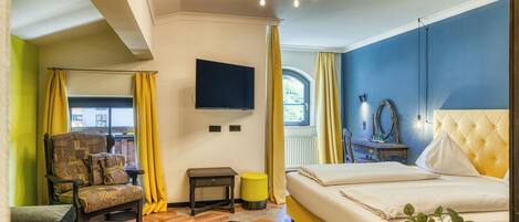 Habitación de hotel con cama, una silla, una mesita, televisión y ventana con cortinas.
