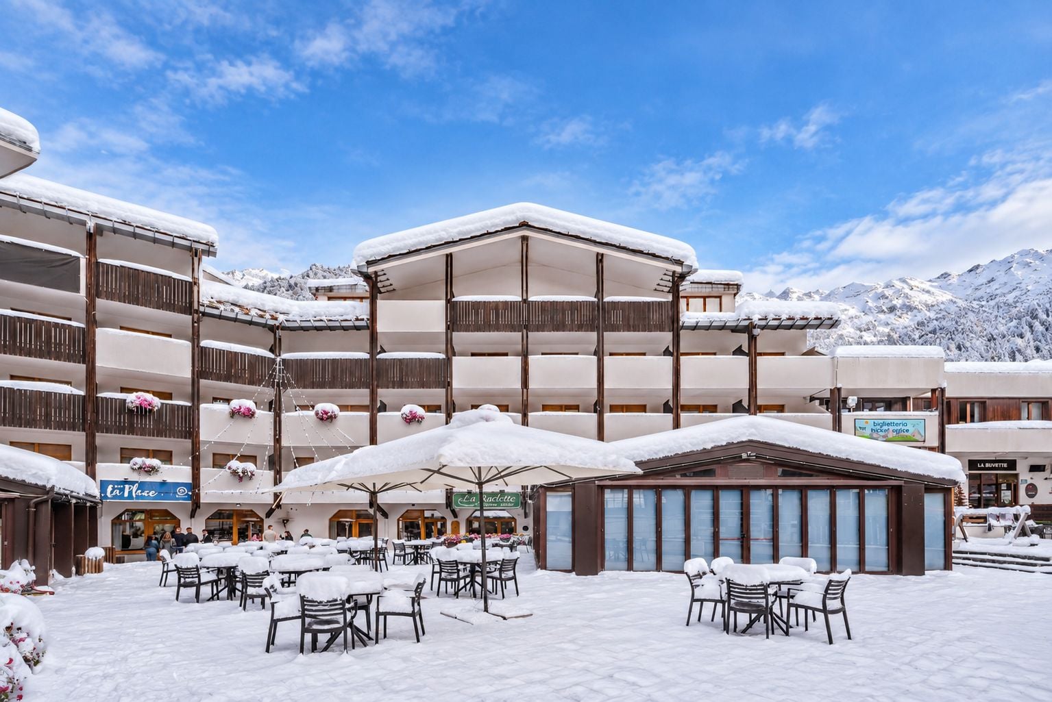 A multi-story hotel with outdoor seating areas covered by snow, surrounded by mountains.