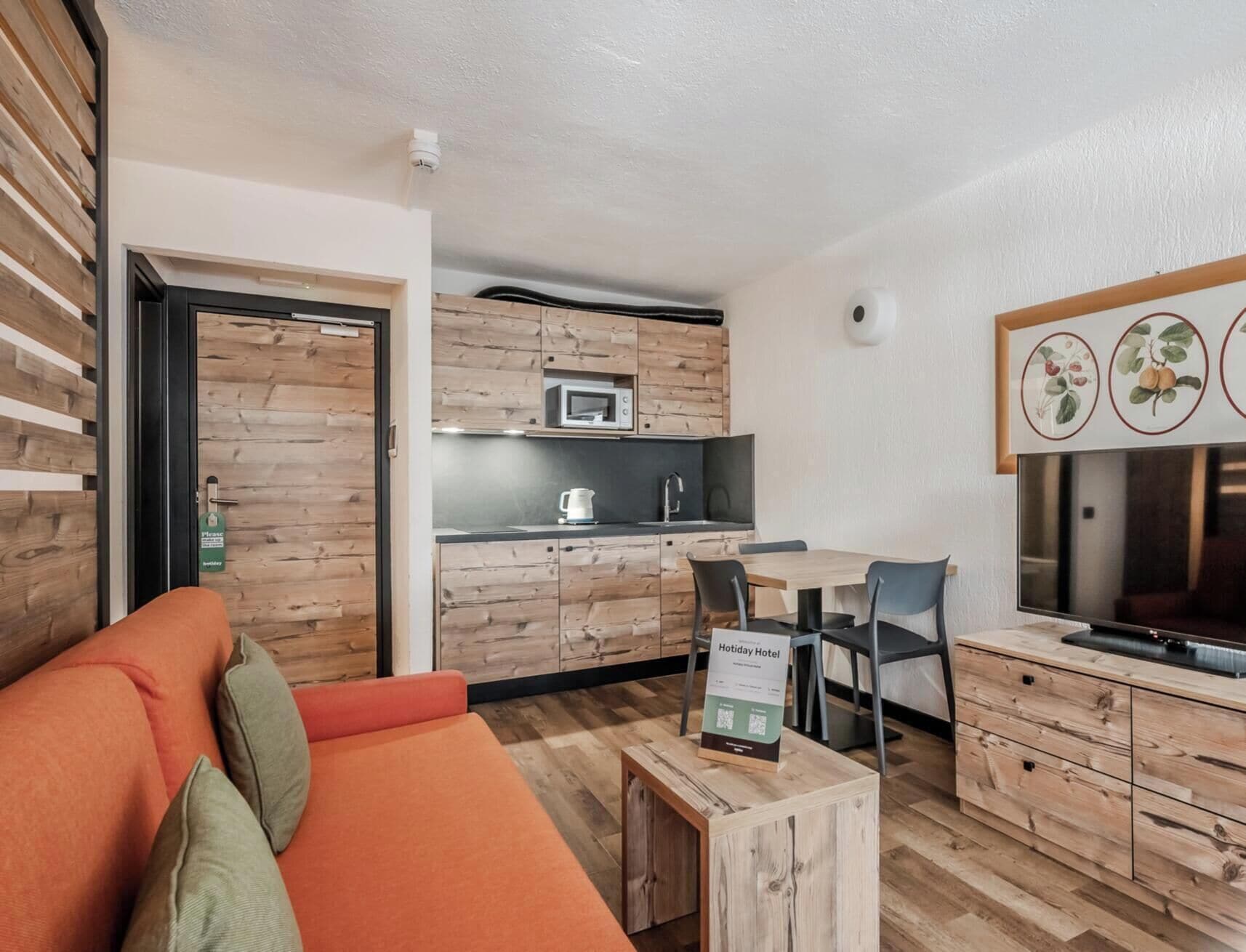 A compact hotel room with a kitchenette, a sofa, a dining area, and a television.