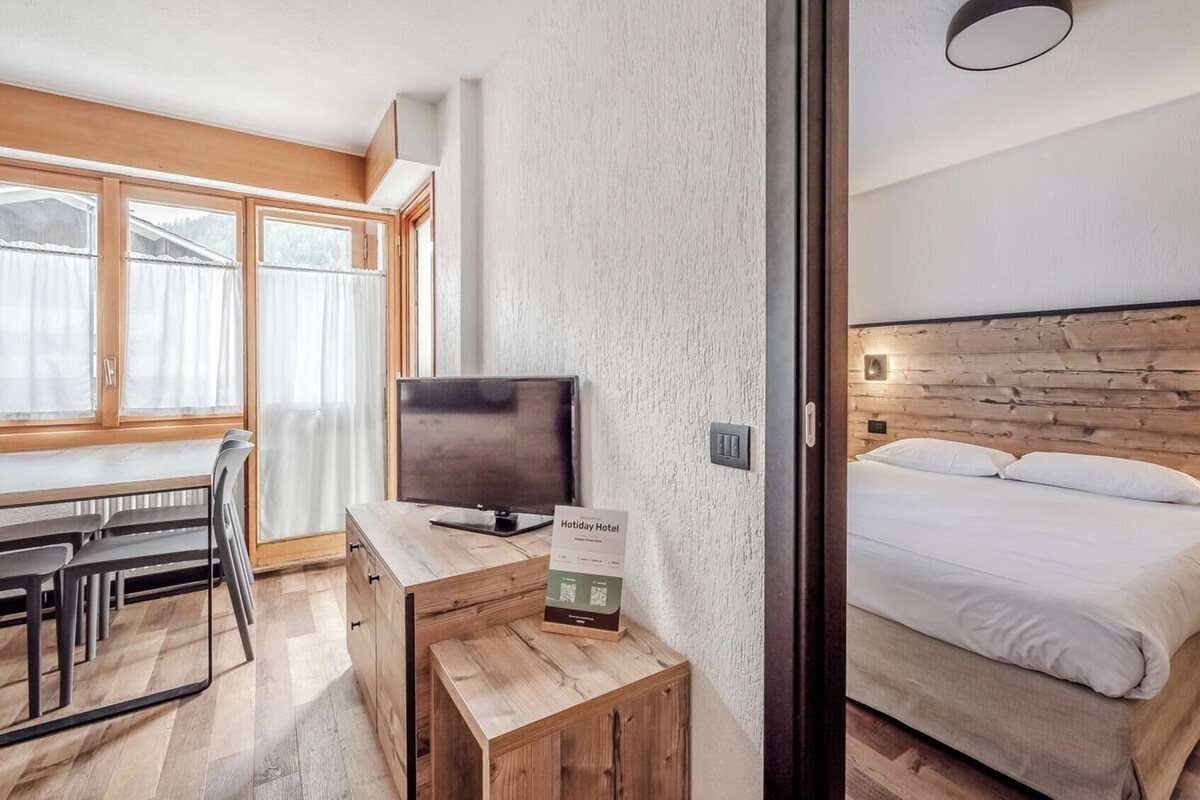 A modern hotel room with a wooden headboard, a bed, a desk with a television, and a small table.