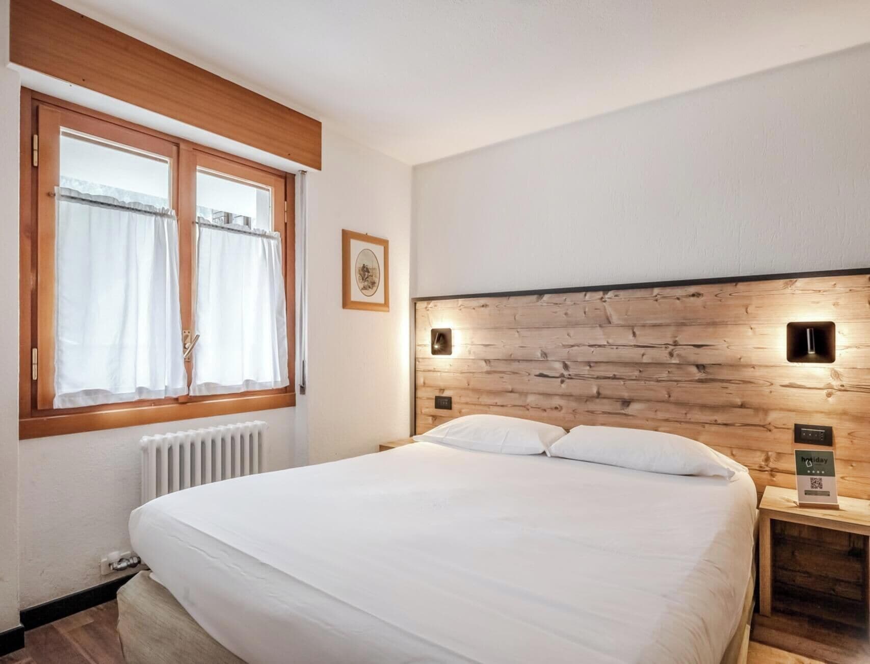 A bedroom with a wooden headboard, a bed with white bedding, a window with curtains, a framed picture on the wall, and a small bedside table with a sign.