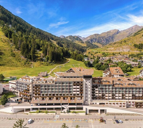 A hotel with multiple floors, a parking area, and a mountainous backdrop. - Hotiday Apartments La Thuile (La Thuile)
