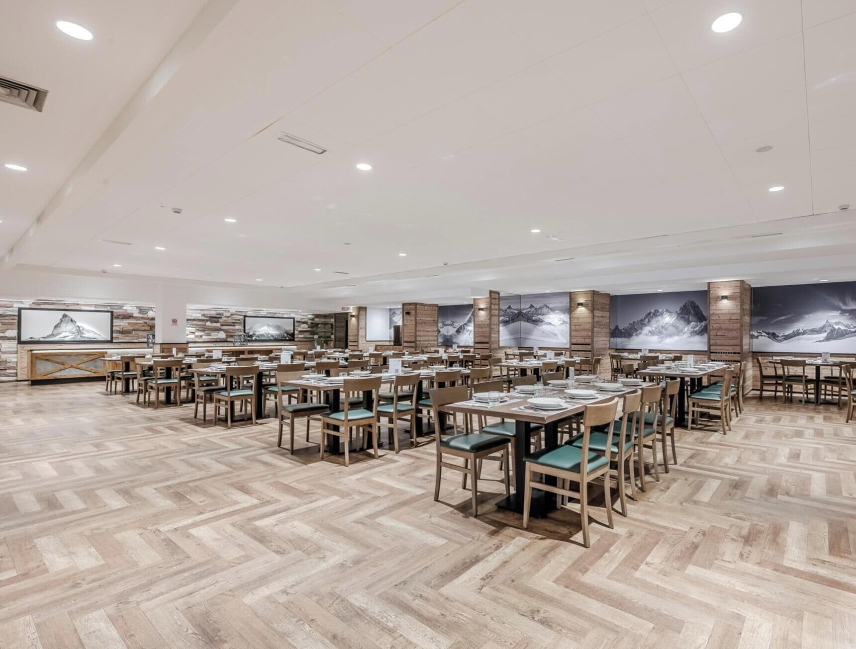 Un comedor amplio con varias mesas y sillas, piso de madera y decoración de temática montañosa.