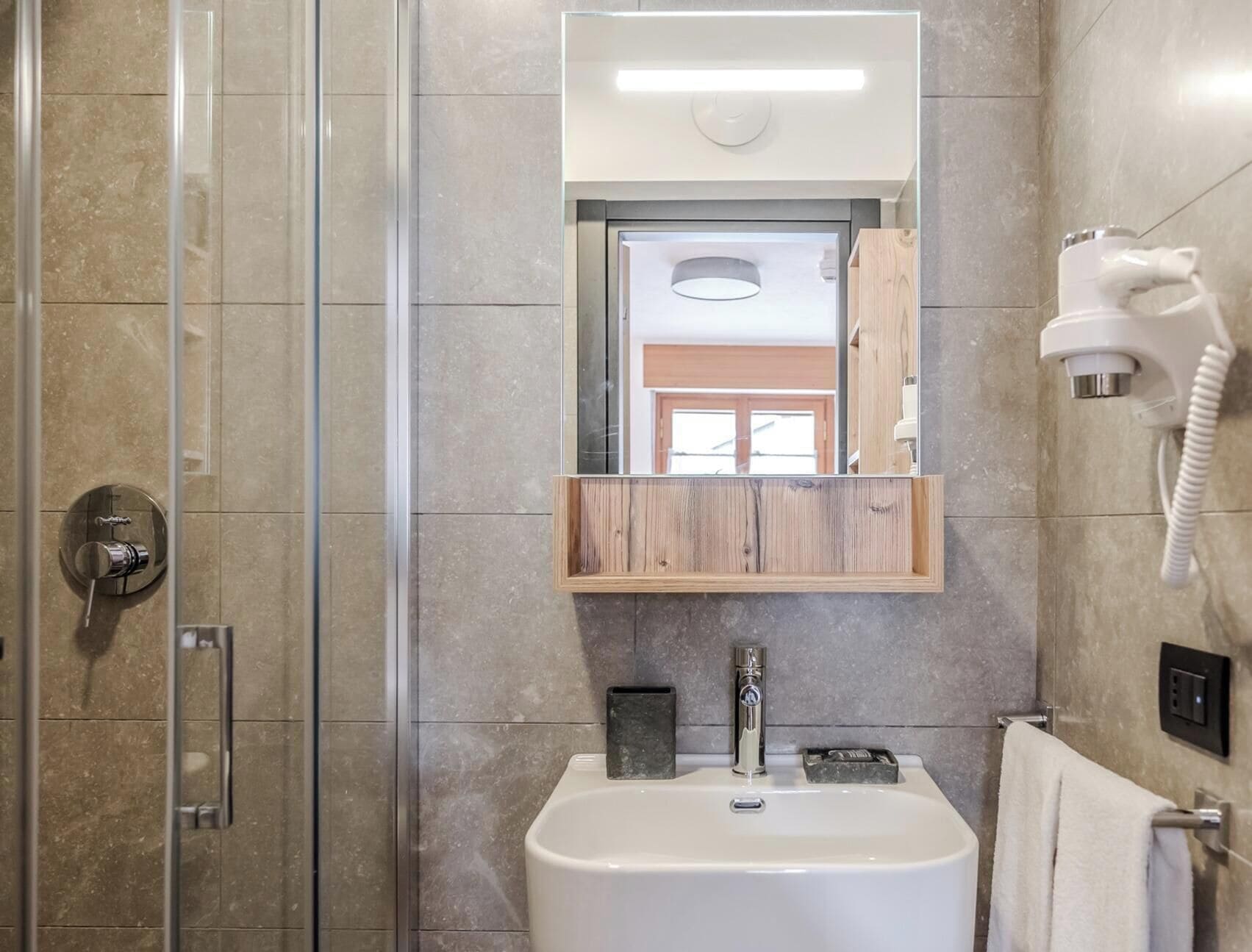 Un baño con ducha, lavamanos, espejo y secador de pelo fijado en la pared.