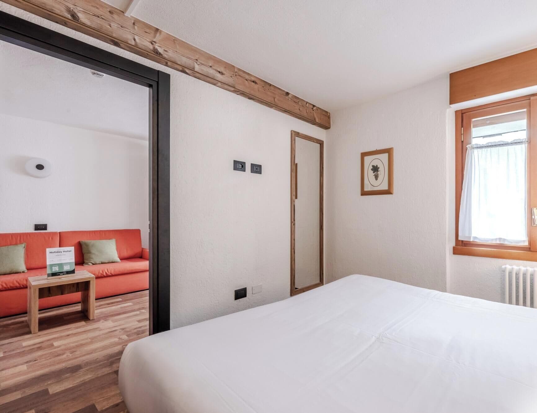 Habitación de hotel con cama, piso de madera, sofá rojo, mesita y ventana con cortinas.