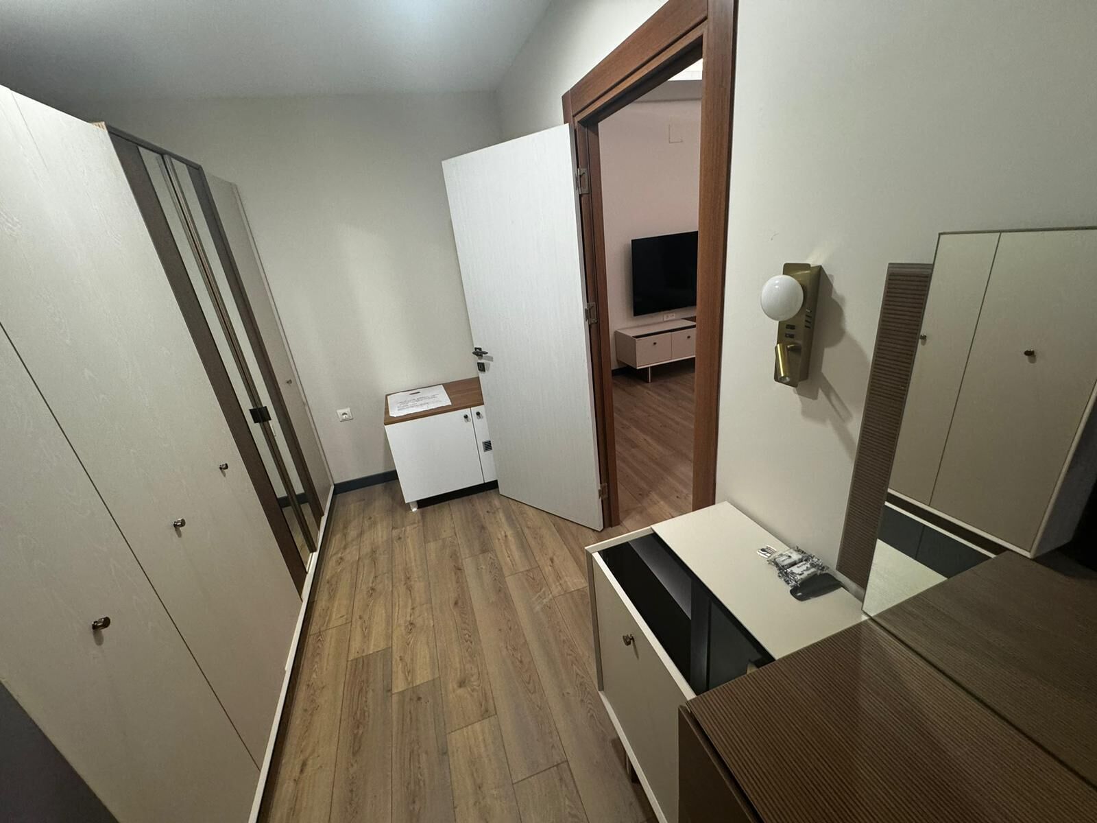 Un couloir étroit avec un plancher en bois, une armoire blanche, un petit bureau avec un miroir et, au fond, un téléviseur.