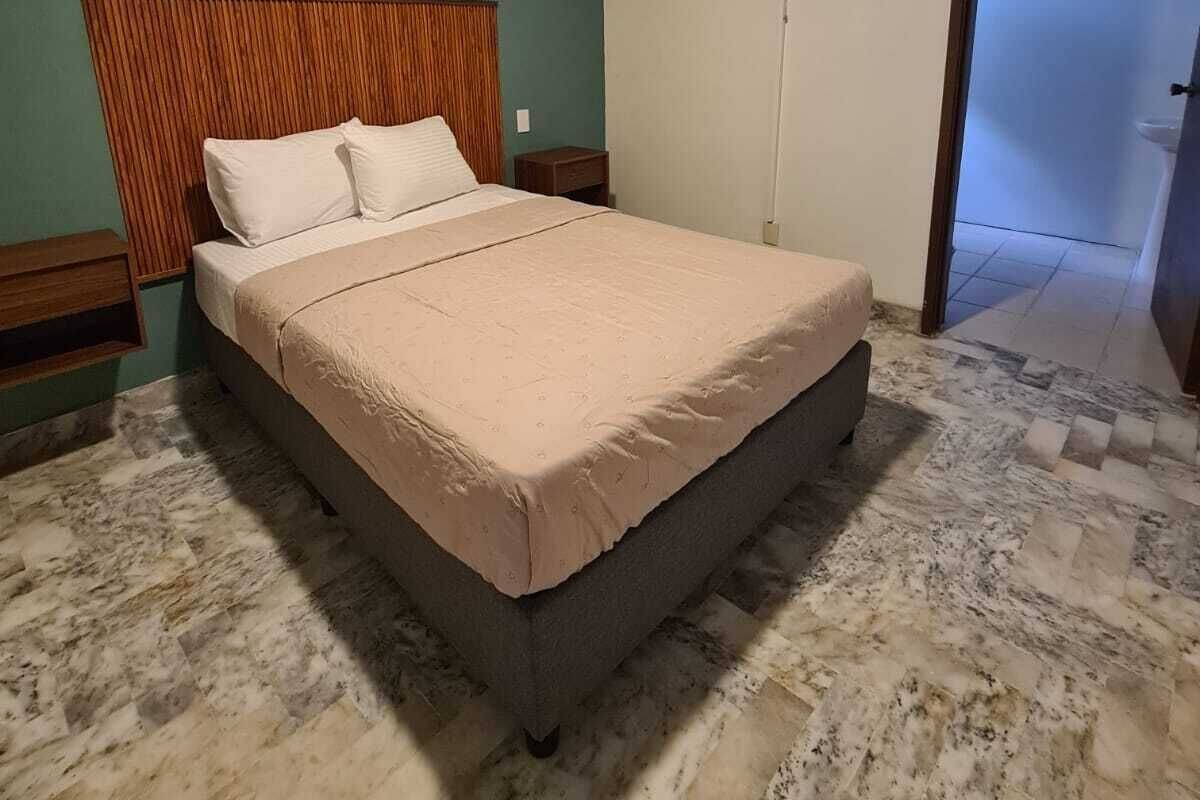 A bedroom with a bed, a wooden headboard, a wall-mounted fan, and a tiled floor.