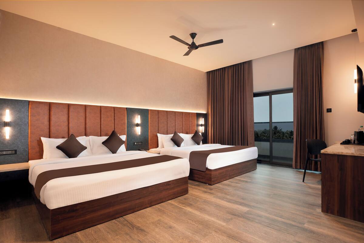 A modern hotel room with two beds, a ceiling fan, a large window with curtains, and a wooden desk.