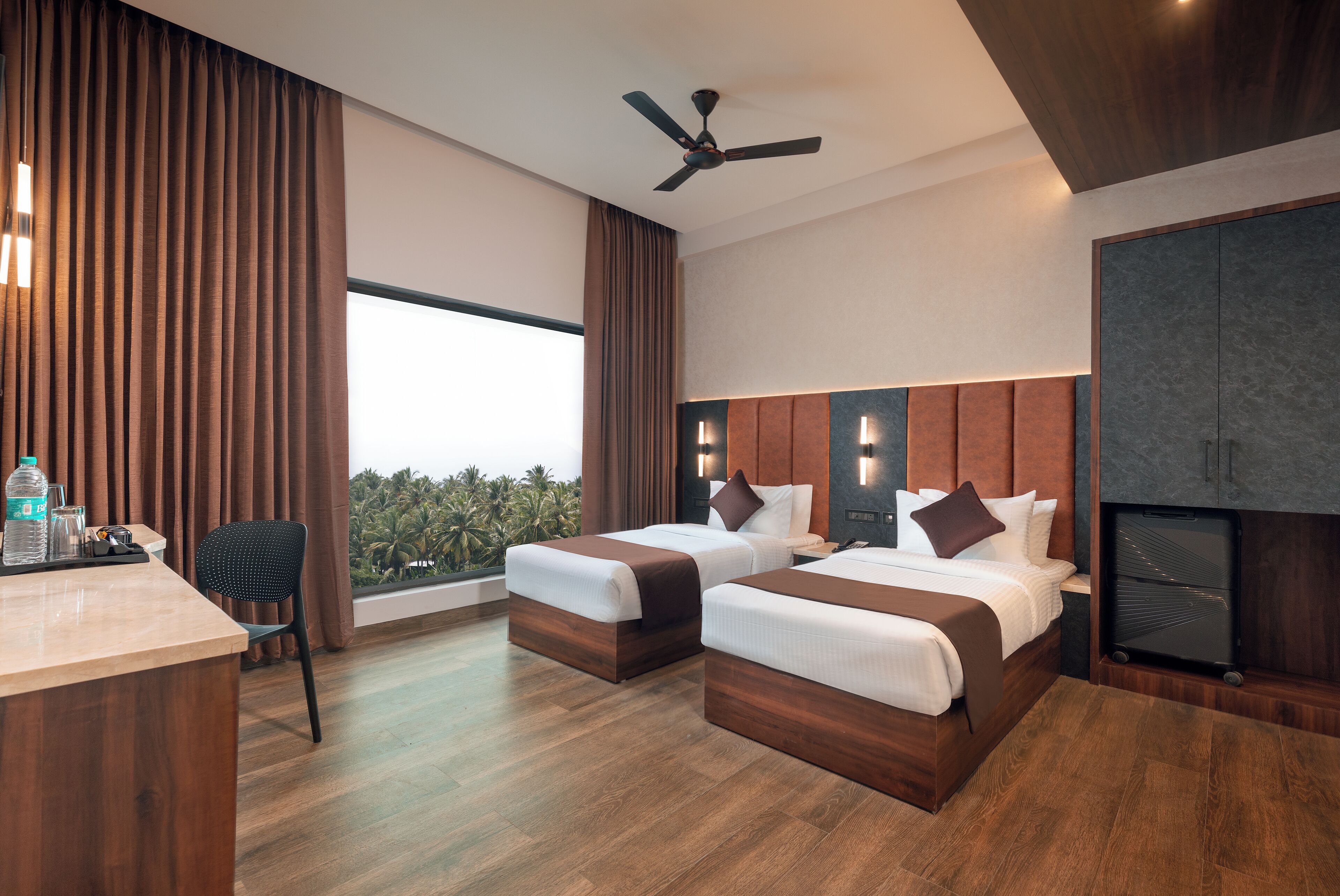 A modern hotel room with two beds, a desk, a chair, a ceiling fan, and a large window with a view of greenery.