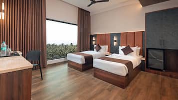 A modern hotel room with two beds, a desk, a chair, a ceiling fan, and a large window with a view of greenery.