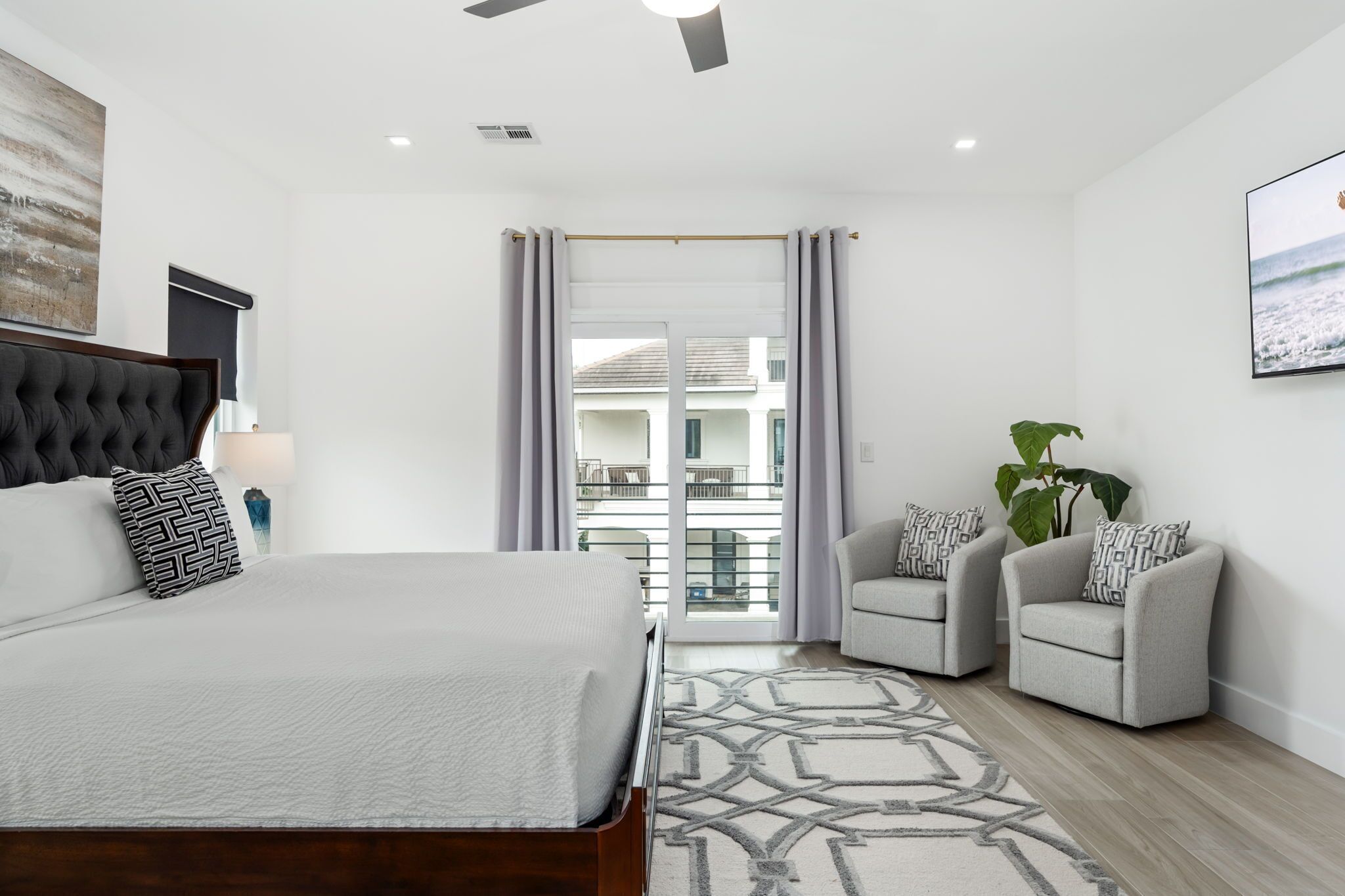 Un dormitorio moderno con una cama grande, dos sillones, un ventilador de techo y vistas al exterior a través de un ventanal.