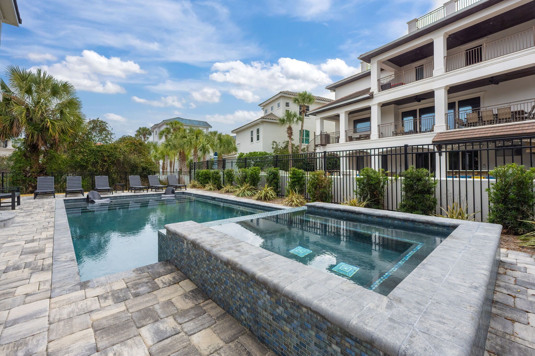 A rectangular swimming pool with a tiled edge, surrounded by a stone patio and a black metal fence.