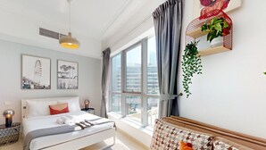 A modern hotel room with a large bed, a bedside table with a lamp, a window with curtains, and a wall-mounted shelf with plants. - Bay Central West - Spacious Studio in the Dubai Marina (Dubai Marina)