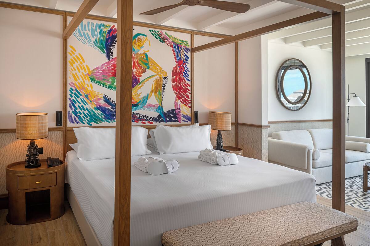 Habitación de hotel con una cama grande, dos mesitas de noche con lámparas, un sofá blanco, un espejo redondo y un cuadro abstracto de colores en la pared.