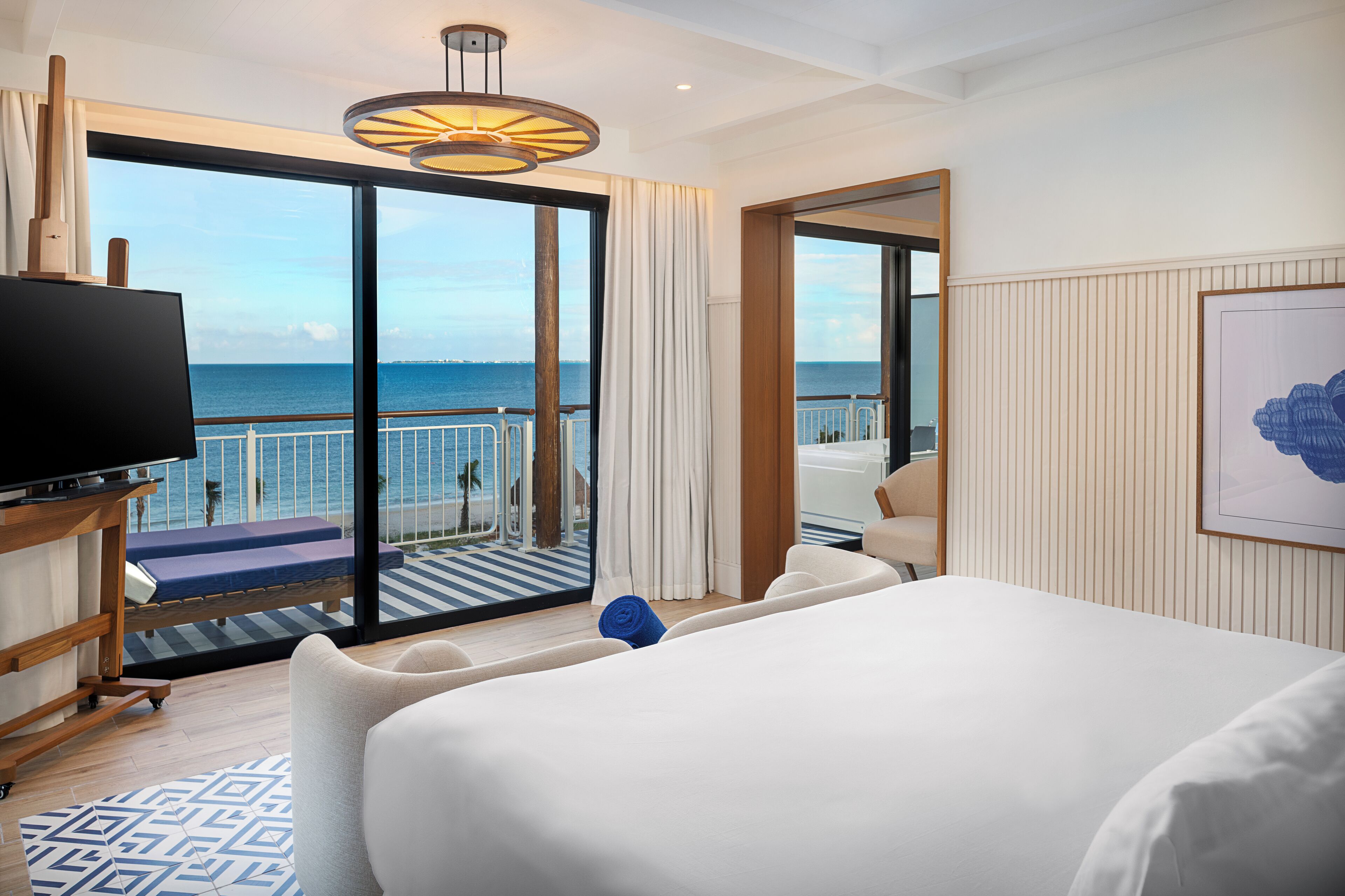 Habitación de hotel con una cama grande, un televisor, un balcón con vista al mar y una obra de arte enmarcada en la pared.