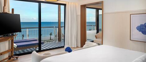 Habitación de hotel con una cama grande, un televisor, un balcón con vista al mar y una obra de arte enmarcada en la pared.